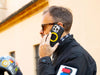Man in sunglasses on a call outdoors holding the MAG case with a yellow ring, helmet resting in the foreground